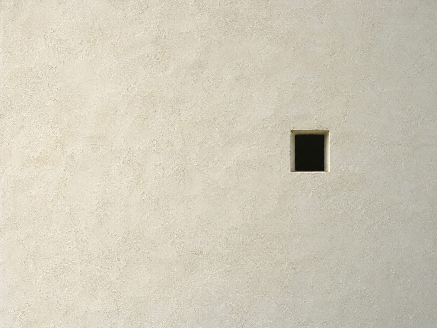 A pale lime-plaster wall filling the frame, with one small square opening set left of centre and no other feature.