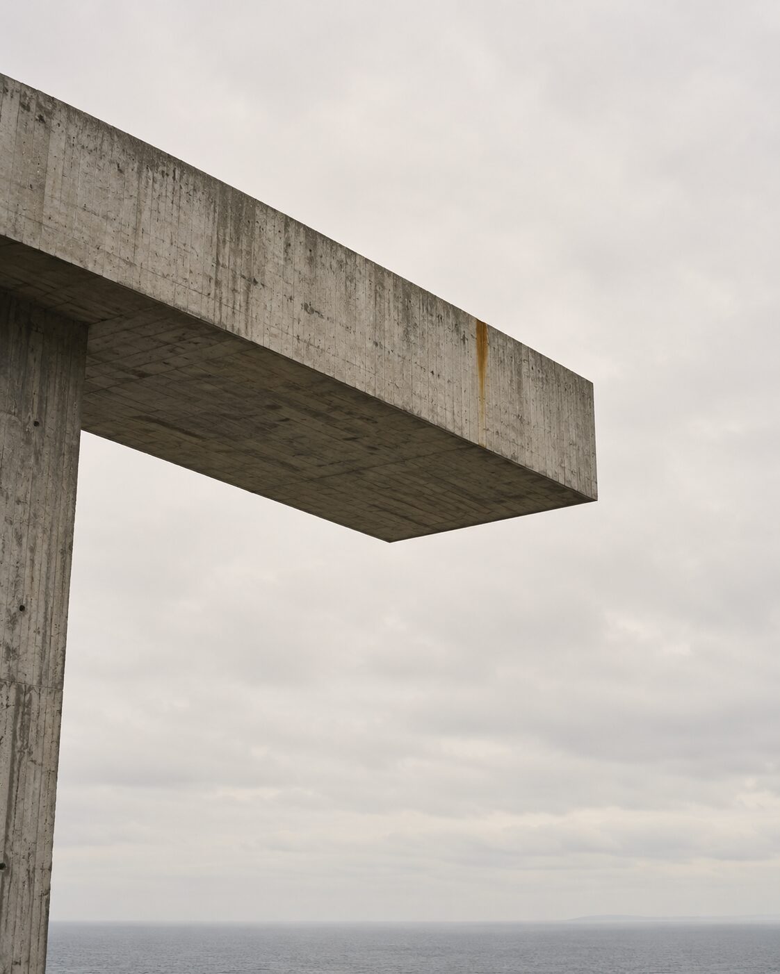 A long concrete slab cantilevered out from a single pier into empty pale sky, seen from below.