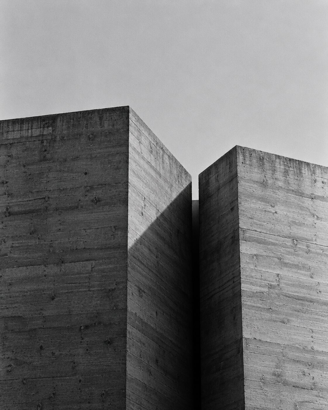 Two board-formed concrete monoliths standing close together, a narrow dark gap between them and one continuous cast shadow.